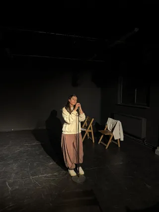 O femeie cu părul lung, purtând un hanorac alb și pantaloni roz, stă pe o scenă întunecată ținând un microfon pe suport.
