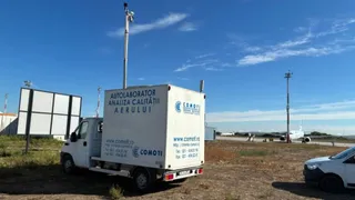 Un autovehicul alb cu un compartiment de marfă marcat cu text și logo este parcat pe un teren de pietriș sub un cer senin.