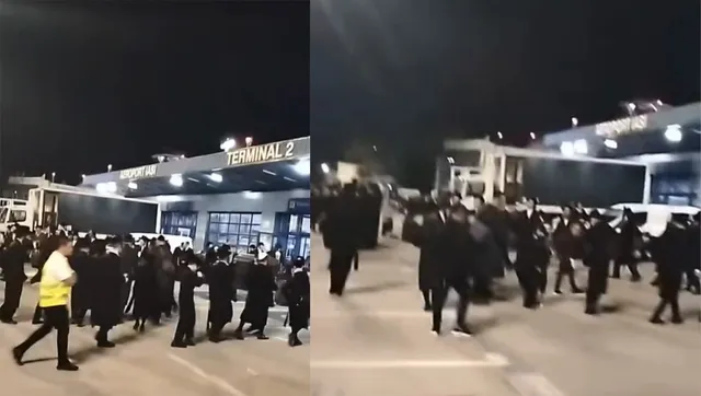 O mulțime de bărbați îmbrăcați în negru, unii cu pălării, se deplasează pe o platformă exterioară a unui aeroport, sub o structură pe care scrie "TERMINAL 2".