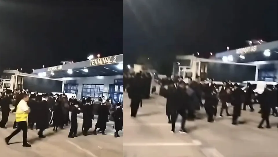 O mulțime de bărbați îmbrăcați în negru, unii cu pălării, se deplasează pe o platformă exterioară a unui aeroport, sub o structură pe care scrie "TERMINAL 2".