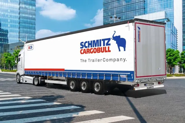 Un camion alb cu remorcă mare, pe care apare un logo albastru cu un elefant și textul Schmitz Cargobull, circulă pe un drum asfaltat.