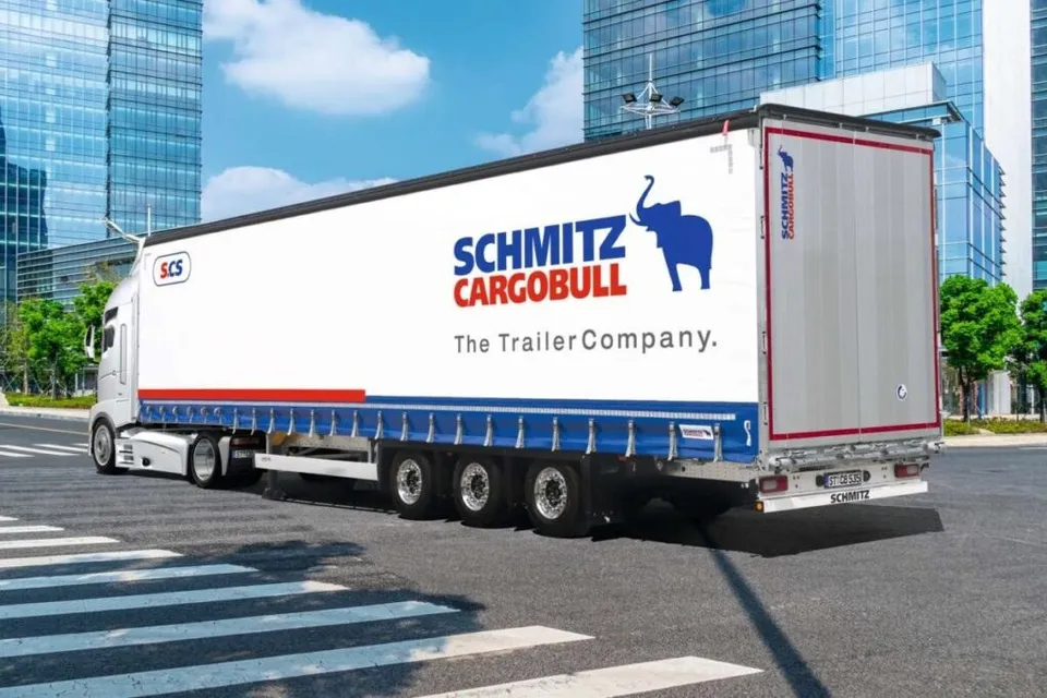 Un camion alb cu remorcă mare, pe care apare un logo albastru cu un elefant și textul Schmitz Cargobull, circulă pe un drum asfaltat.