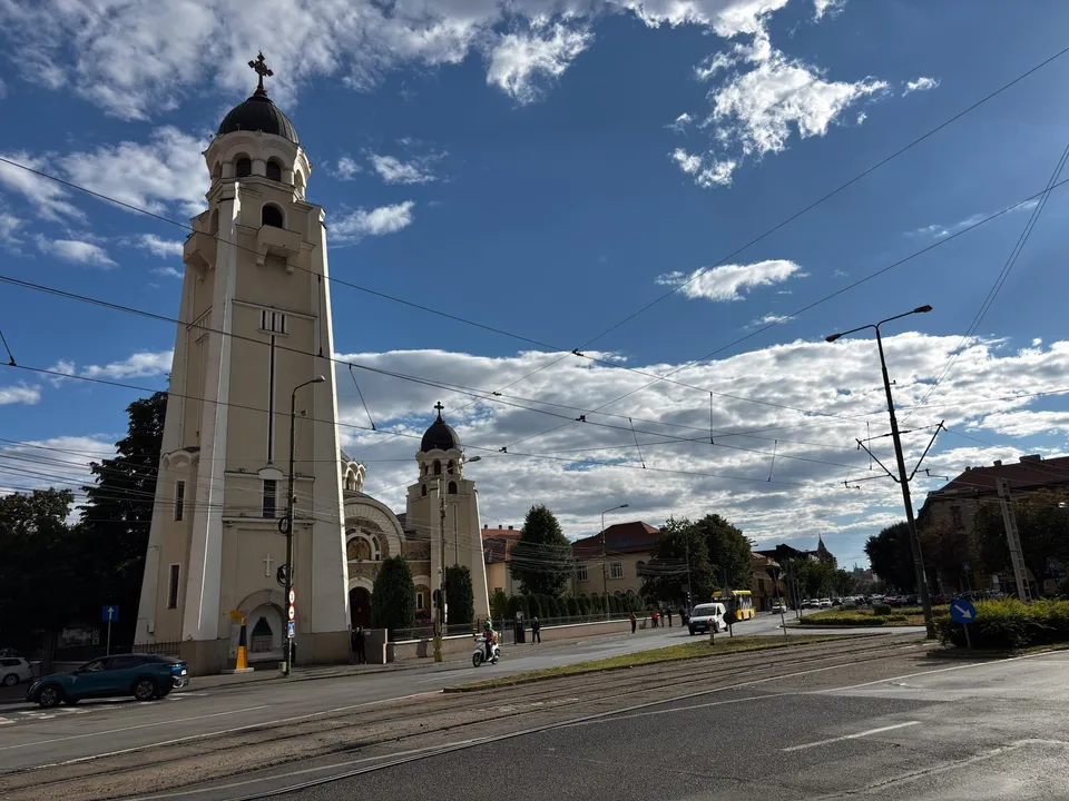 Un turn de biserică înalt și o clădire cu cupolă se ridică lângă o stradă cu șine de tramvai sub un cer cu nori.