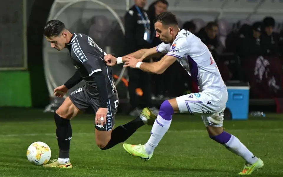 Un jucător în echipament negru și unul în echipament alb se confruntă pe un teren de fotbal cu un balon pe iarbă.