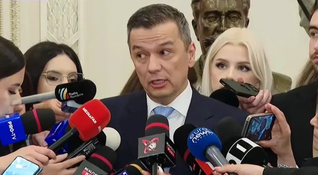 Un bărbat într-un costum închis și cămașă albă este înconjurat de microfoane și persoane care îi îndreptă dispozitive de înregistrare spre față.