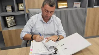 Un bărbat cu cămașă deschisă la guler scrie într-un dosar deschis pe un birou de lemn.
