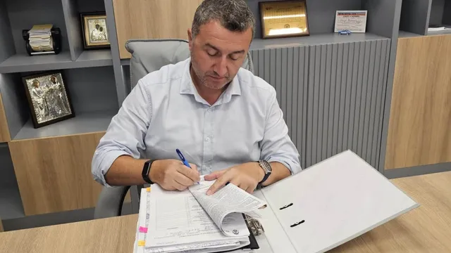Un bărbat cu cămașă deschisă la guler scrie într-un dosar deschis pe un birou de lemn.