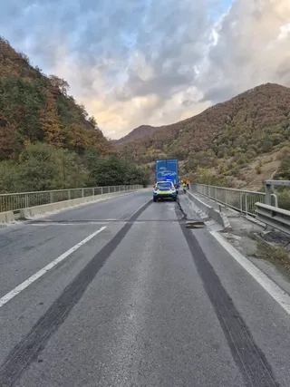 Un vehicul de poliție cu luminile aprinse stă pe o porțiune de drum asfaltat, înconjurat de dealuri împădurite sub un cer înnorat.