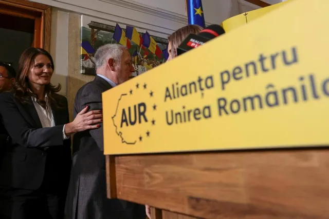 O femeie într-un costum negru pune mâna pe spatele unui bărbat în costum întunecat lângă un podium galben cu text scris.