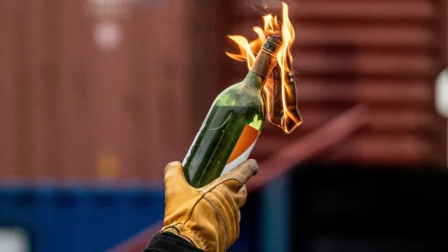 O mână cu mănușă de protecție maro ține o sticlă de sticlă verde cu un obiect aprins în flăcări în partea superioară.