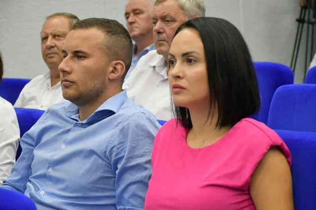 Un bărbat cu cămașă albastră și o femeie cu bluză roz stau așezați în linii, cu alți bărbați în fundal.