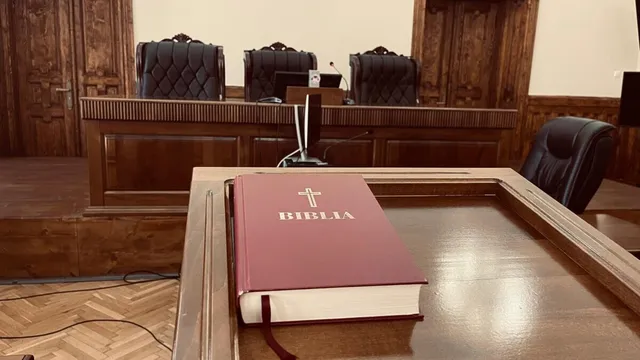 O Bible roșie cu o cruce și textul alb se află pe o masă de lemn în fața unor scaune de birou negre.