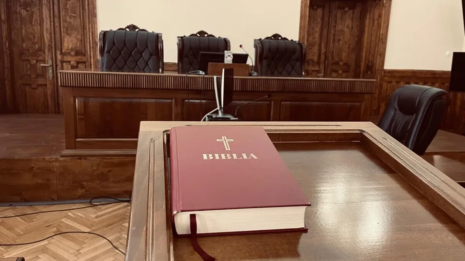 O Bible roșie cu o cruce și textul alb se află pe o masă de lemn în fața unor scaune de birou negre.