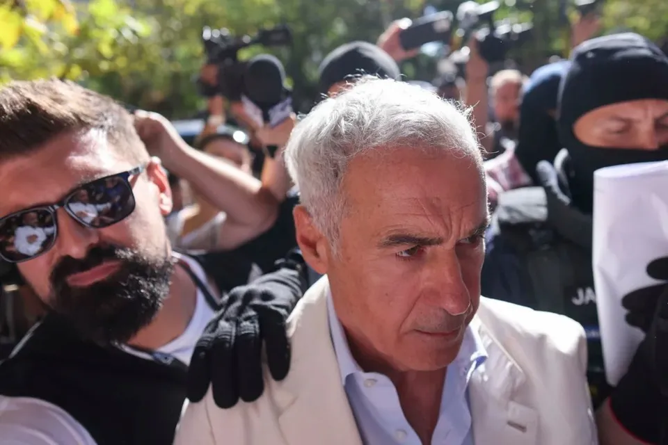 Un bărbat cu părul alb și sacou deschis este înconjurat de persoane, dintre care una poartă ochelari de soare și o altă persoană poartă o mască neagră.