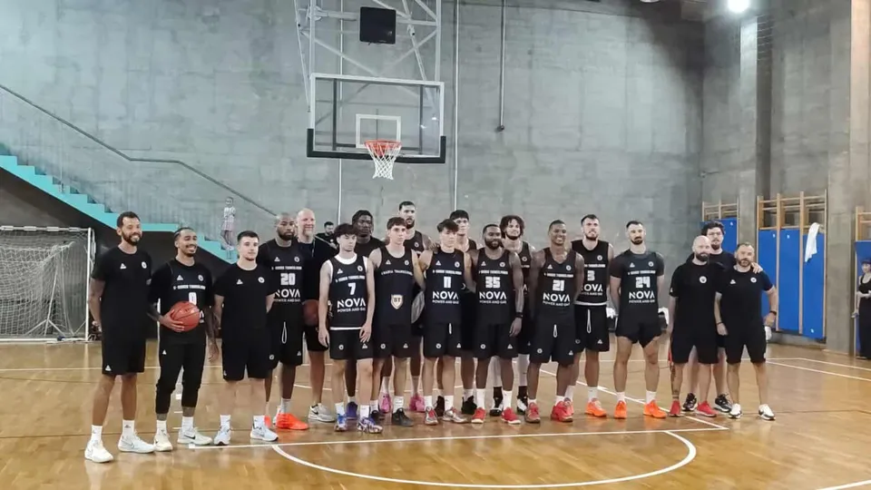 Un grup de sportivi în echipamente negre stau în rând pe un teren de baschet interioară sub un panou de baschet suspendat.