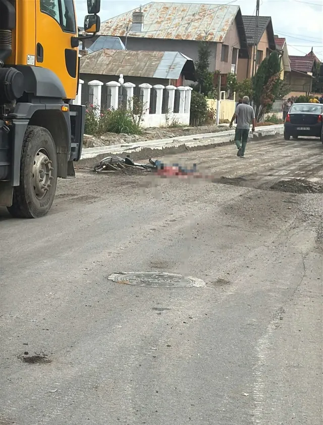 O autoutilitară galbenă este parcată pe marginea unui drum, lângă o bicicletă și o pată roșie pe asfalt, în timp ce un bărbat merge pe trotuar.