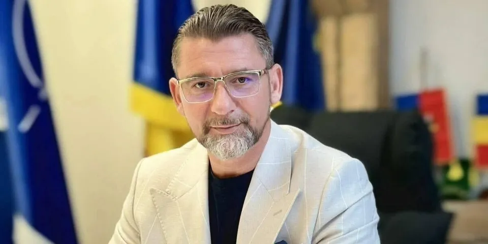 Un bărbat cu ochelari, barbă scurtă și sacou deschis stă în fața unor steaguri albastre și galbene.