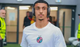 Un tânăr bărbat cu păr scurt, mustăți și urechi mari purtând căști mari de tip over-ear, într-o tricou alb cu un logo rotund multicolor pe piept.