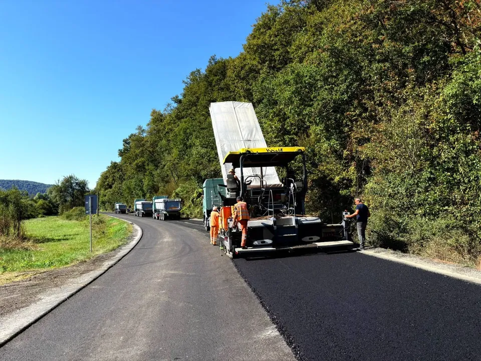 O mașină de asfalt și un camion de transport lucrează pe un drum asfaltat lângă o zonă cu vegetație densă sub un cer senin.