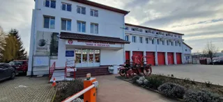 O clădire albă cu ferestre multiple este situată lângă un vehicul de pompieri de epocă și o barieră porti-culoare portocalie.