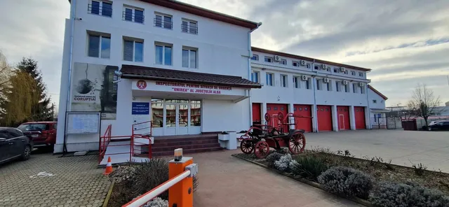 O clădire albă cu ferestre multiple este situată lângă un vehicul de pompieri de epocă și o barieră porti-culoare portocalie.