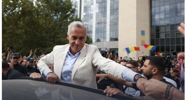 Un bărbat cu părul alb și sacou deschis stă într-un vehicul, întinzând mâna spre o mulțime de oameni cu telefoane și aparate foto.
