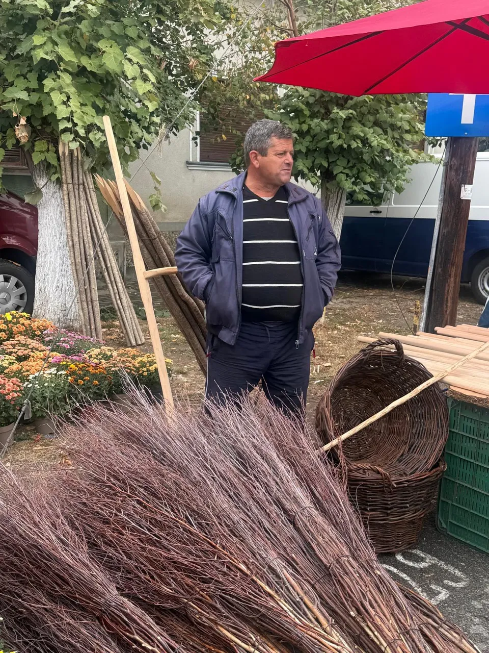 A man in a blue jacket and striped shirt stands behind bundles of brooms and woven baskets at an outdoor market.