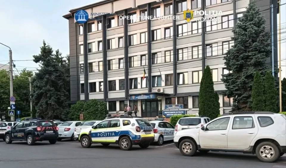 Un vehicul de poliție albastru și alb este parcat în fața unei clădiri înalte de birouri lângă alte mașini albe și negre.