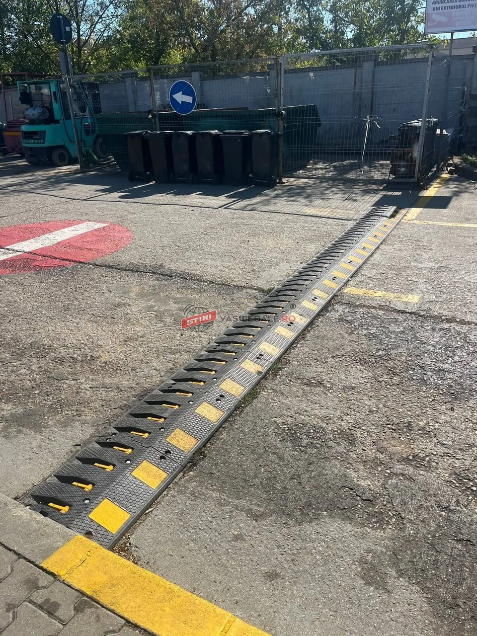 Un dispozitiv de tip bumper, de culoare neagră cu dungi galbene, este montat pe o suprafață asfaltată lângă un gard metalic și containere.
