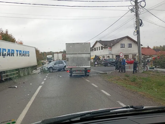 Un camion mare și un autoturism gri sunt poziționate pe un drum asfaltat lângă un grup de persoane și alte vehicule parcate.