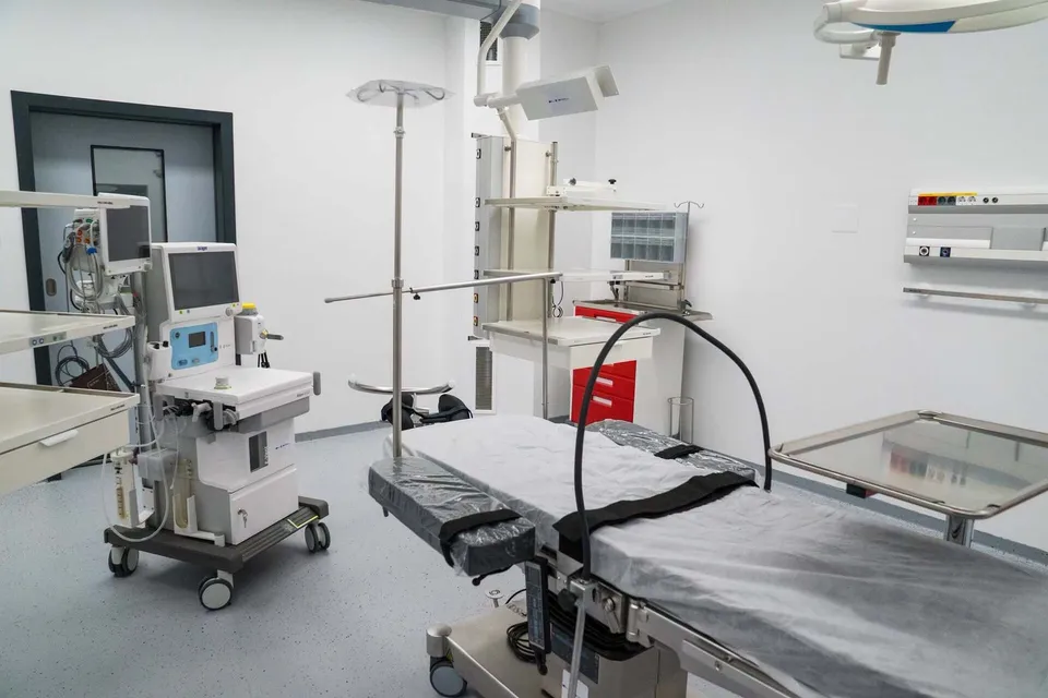 O cameră medicală cu un pat de spital, un dispozitiv monitorizat pe un suport cu roți și echipamente medicale pe pereți și mese albe.