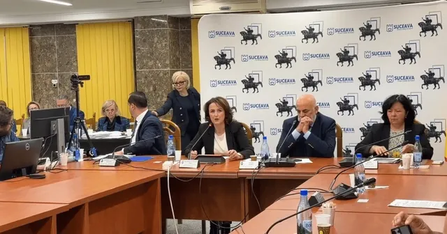 Oameni stau la o masă lungă de conferințe într-o sală cu un fundal alb decorat cu logouri repetate, având computere și sticle de apă în față.