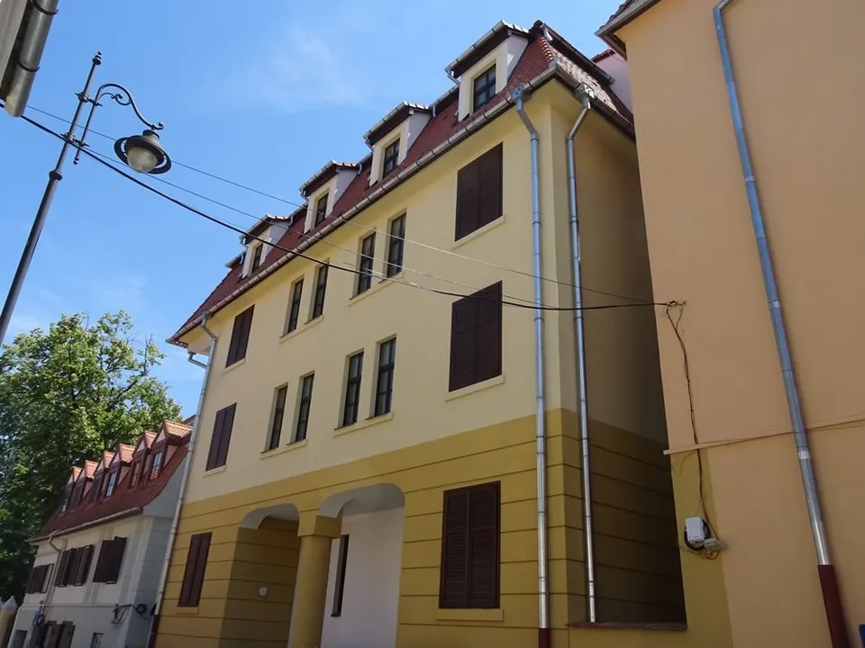 O clădire cu fațadă galbenă și acoperiș înclinat dispune de ferestre cu obloane maronii și țevi de scurgere metalice pe perete.