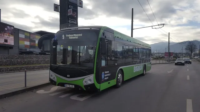 Un autobuz verde și negru cu numărul 9 circulă pe un drum asfaltat lângă un trotuar și un panou publicitar înalt.