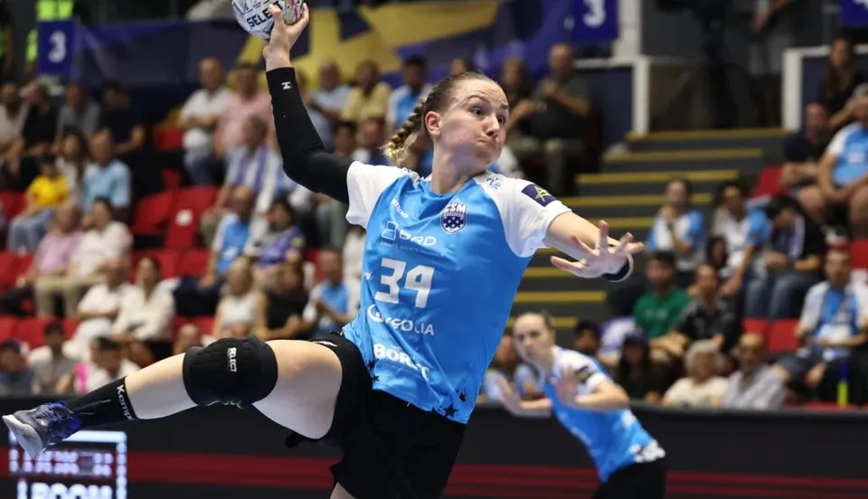 O jucătoare de handbal în echipă albastră și neagră aruncă mingea într-o poziție suspendată pe un teren de sport cu spectatori în fundal.