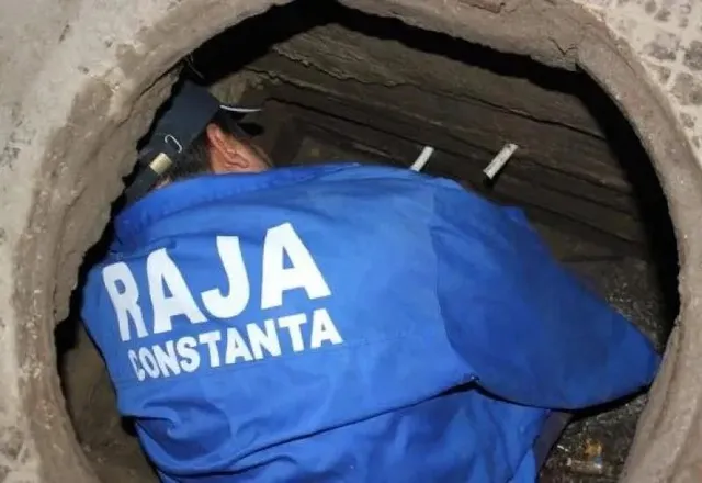 Un bărbat cu o șapcă și o haină albastră pe care scrie RAJA CONSTANTA este aplecat într-o conductă sau un șanț întunecat.