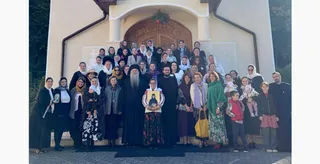Un grup de persoane, inclusiv femei cu batic și bărbați în haine negre, stau grupate în fața unei clădiri cu un arc de intrare.