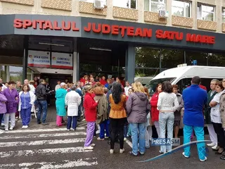 Un grup de persoane stă în fața unei clădiri cu inscripția SPITALUL JUDEȚEAN SATU MARE, lângă o ambulanță albă.