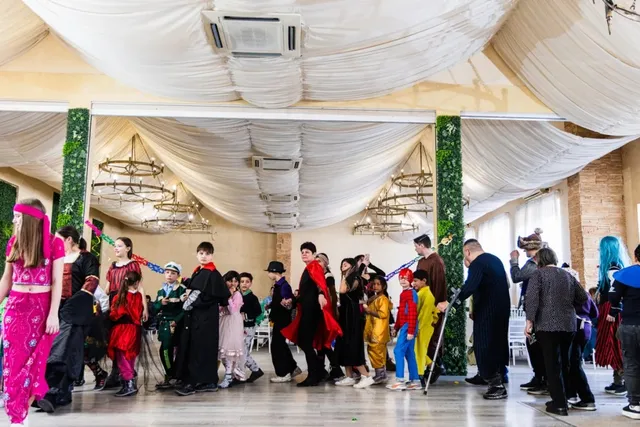 Un grup de persoane de diverse vârste, îmbrăcate în costume colorate și diverse haine, stau într-o încăpere decorată cu draperii albe și lumini suspendate.