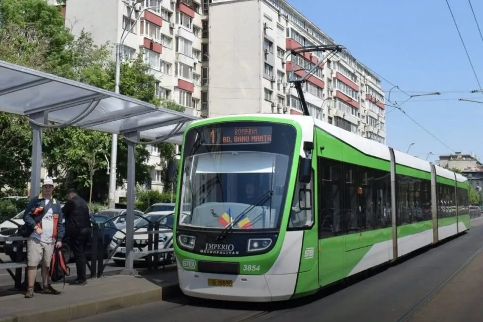 Un tramvai verde și alb circulă pe o șine lângă o stație de autobuz unde stau doi bărbați în fața unui bloc de locuințe.
