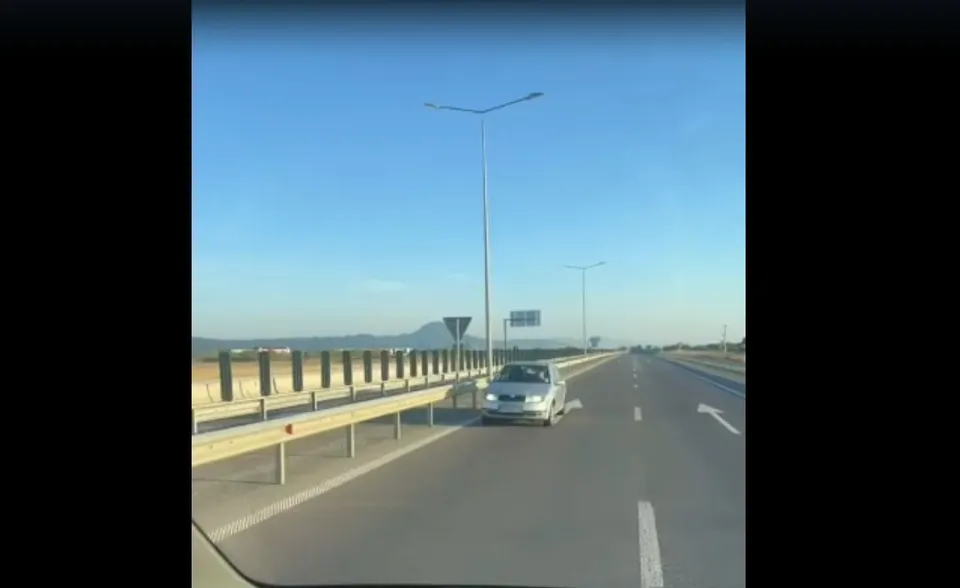 O mașină de culoare albă circulă pe o autostradă cu marcaje rutiere și stâlpi de iluminat sub un cer senin.