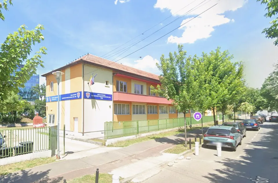 O clădire cu fațadă galbenă și albă este situată lângă un drum cu mașini parcate și copaci verzi sub un cer înzăpezit.