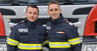 Doi bărbați în uniforme de pompieri cu benzi reflectorizante galbene stau în fața unui vehicul alb.