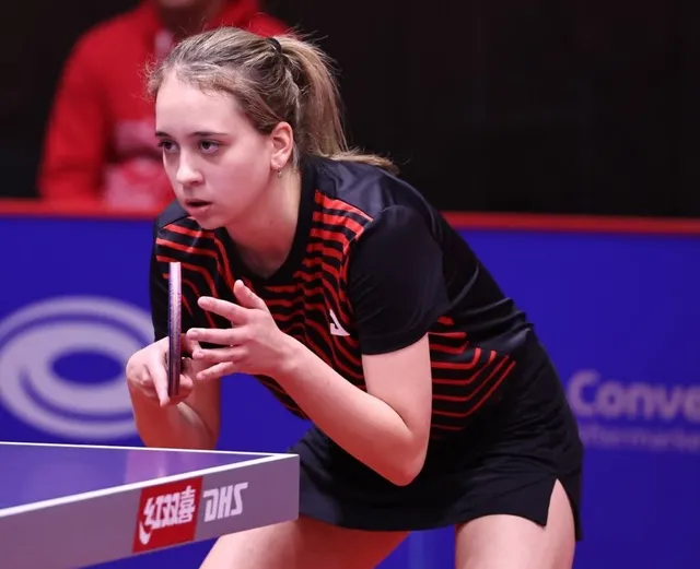 O tânără sportivă cu părul legat într-o coadă de cal ține o paletă de tenis de masă în poziție de pregătire lângă o masă.