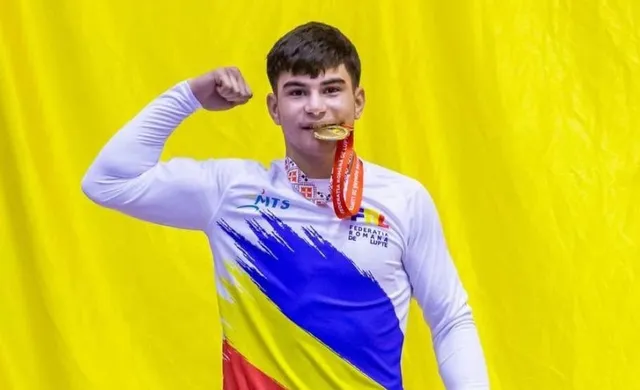 Un bărbat cu tricou alb cu modele colorate poartă o medală aurie în jurul gâtului și ridică pumnul în fața unui fundal galben.