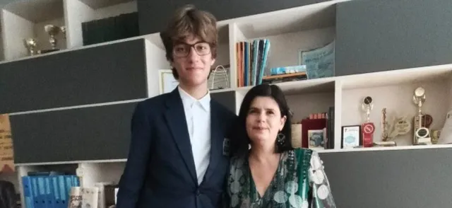 Un bărbat tânăr cu ochelari și o femeie stau în fața unor rafturi cu cărți și trofee într-o încăpere interioară.
