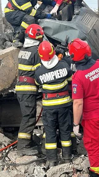 Patru persoane îmbrăcate în uniforme de salvator, unele cu cască roșie și inscripții, stau lângă un vehicul gri și resturi de beton.