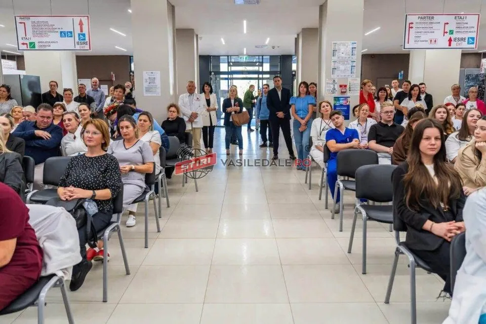 Oamenii stau așezați pe scaune negre într-un hol cu podea deschisă și pereți albi, privind spre centrul încăperii.