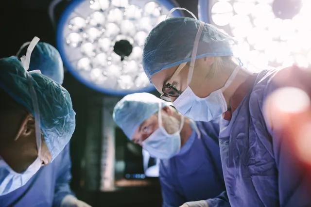 Trei medici cu măști și bonete de protecție albastre stau deasupra unei mese de operație sub lumini puternice de spital.