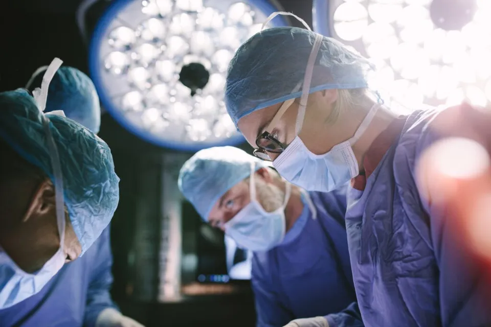 Trei medici cu măști și bonete de protecție albastre stau deasupra unei mese de operație sub lumini puternice de spital.